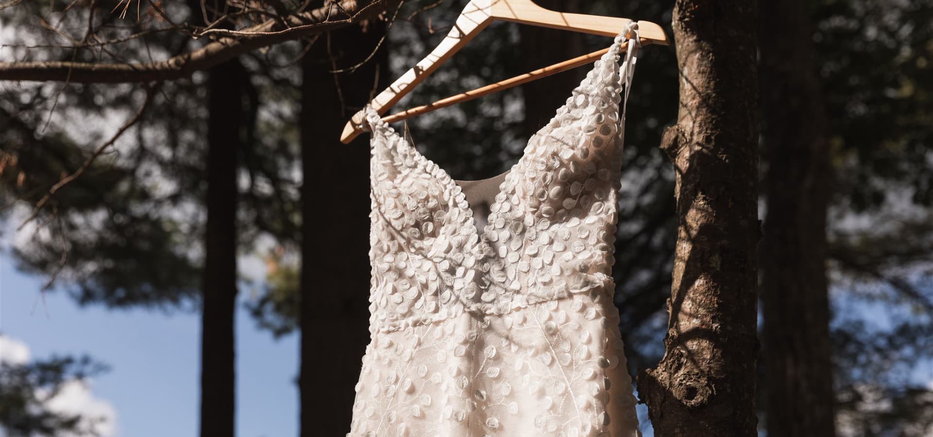 A delicate white dress with floral details hangs from a wooden hanger among the trees.