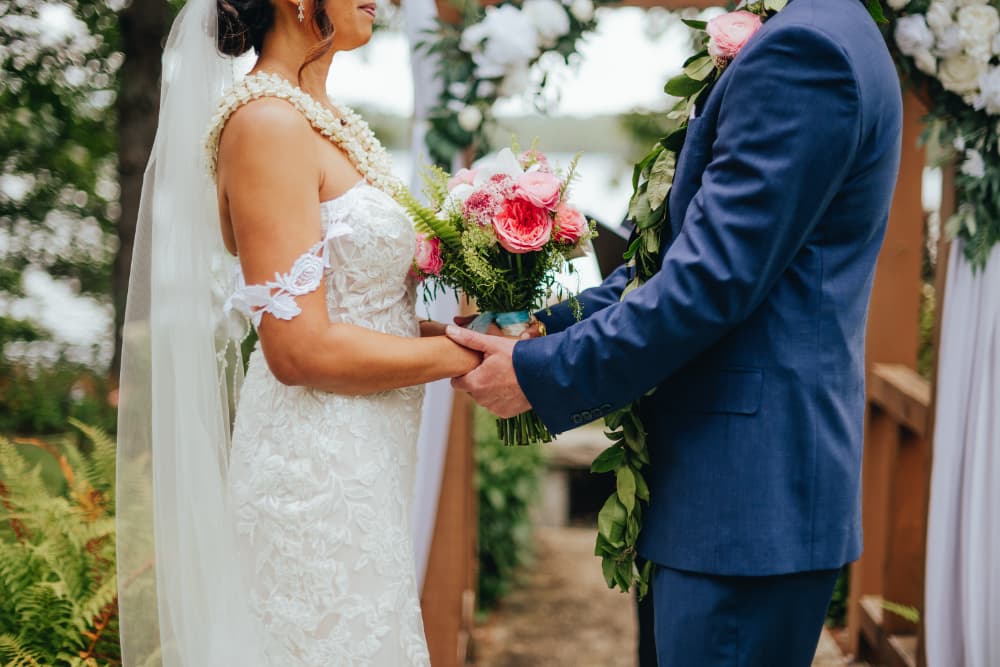 A bride and groom hold hands during their wedding ceremony, surrounded by flowers and greenery.