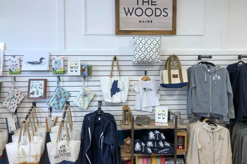 A retail display featuring clothing and merchandise with nature-themed designs and the name "The Woods, Maine."