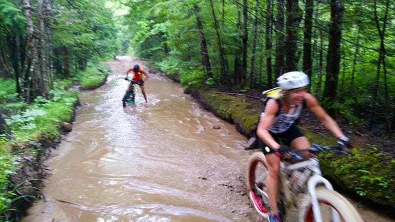 Two cyclists navigating a muddy trail surrounded by dense vegetation.