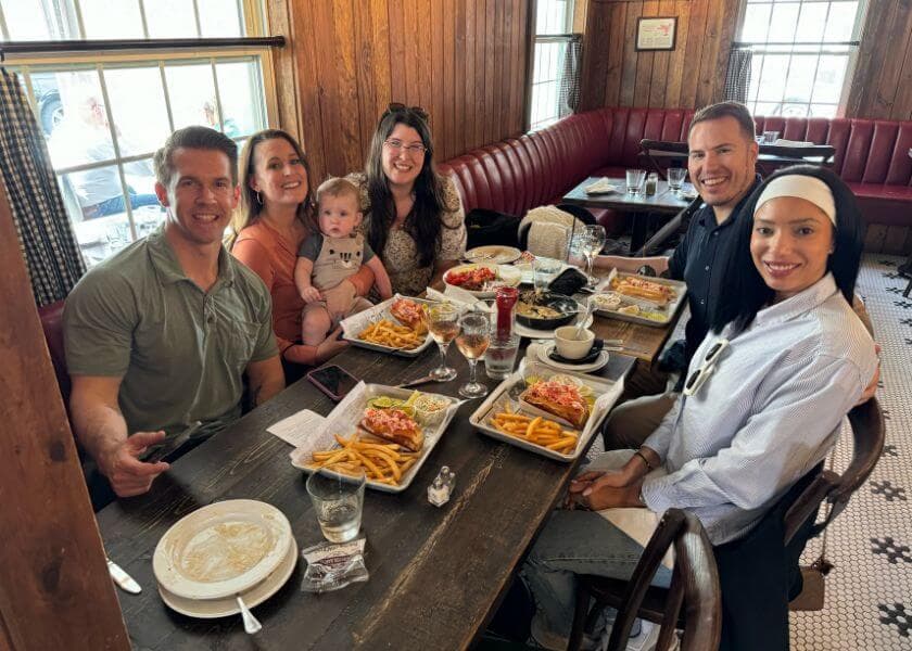 A group of six adults and a baby enjoy a meal together at a wooden table in a cozy restaurant.