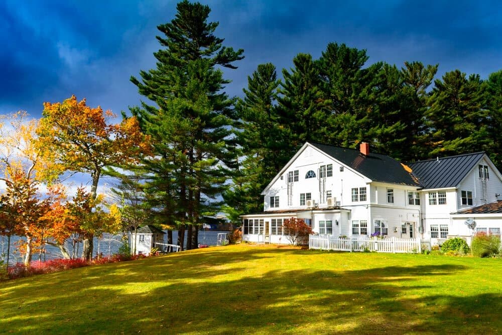 A white house with a dark roof surrounded by tall green trees and autumn foliage under a dramatic sky.