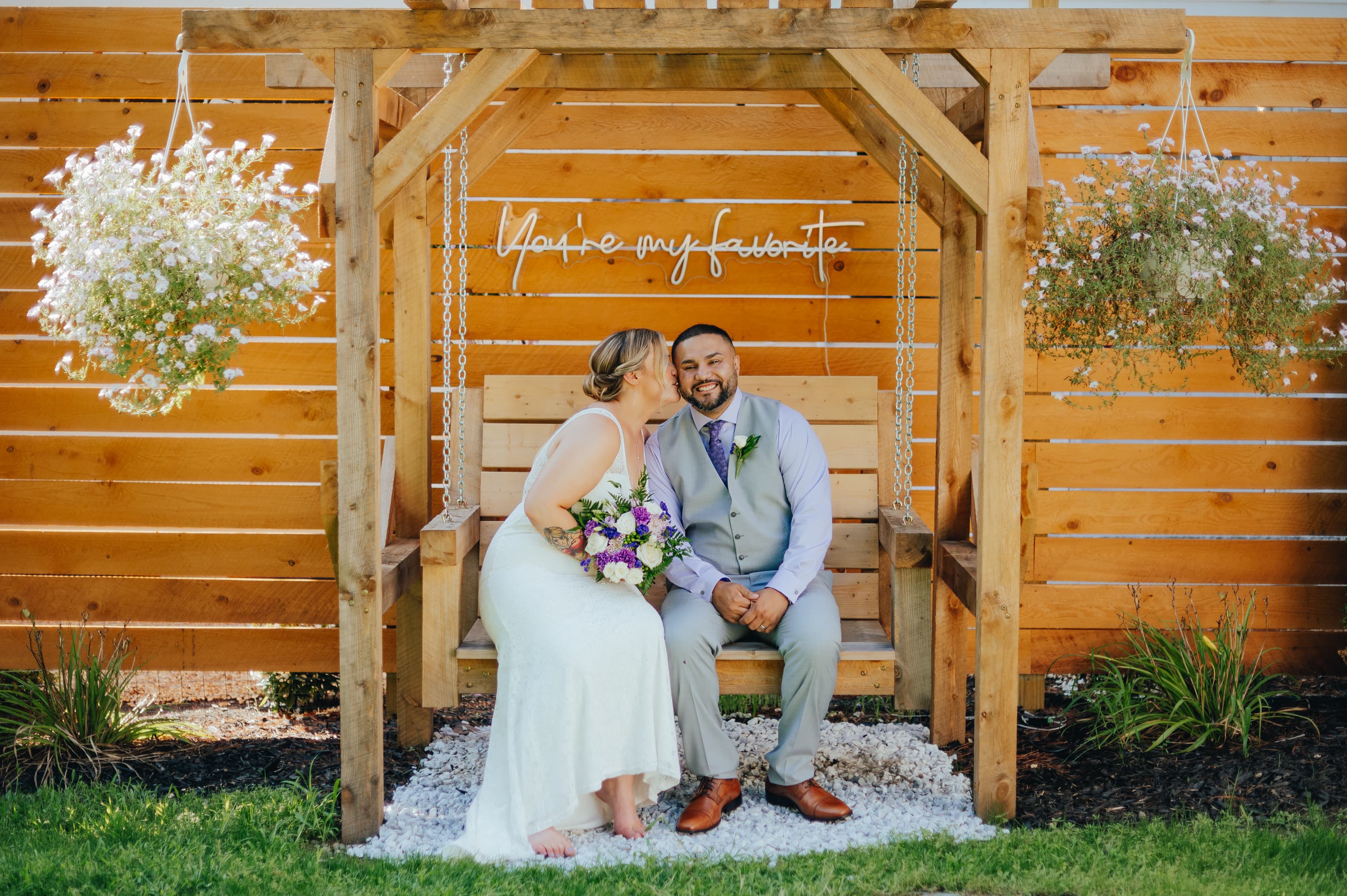 A couple shares a joyful moment on a swing under a wooden gazebo, surrounded by flowers and a wooden backdrop.