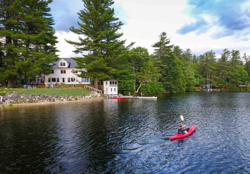 A person kayaks on a calm lake near a house surrounded by tall trees.