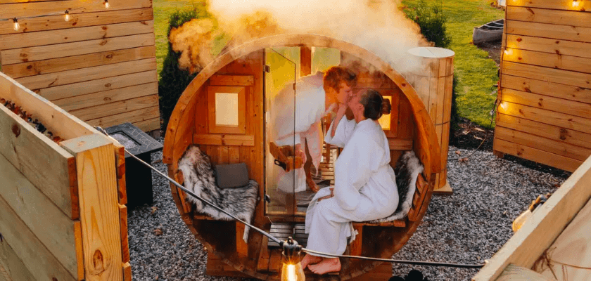 Two women in bathrobes kiss inside a wooden sauna, surrounded by a cozy outdoor setting.