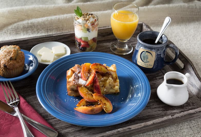 A wooden tray holds a blue plate featuring baked apple dish, alongside a muffin, butter, fruit parfait, orange juice, and coffee.