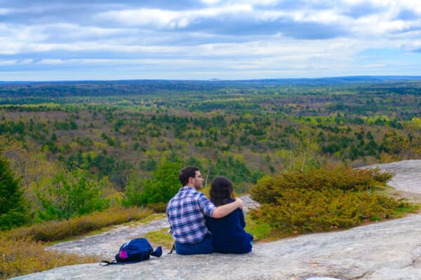 A couple sits together on a rock, overlooking a vast landscape of trees under a cloudy sky.
