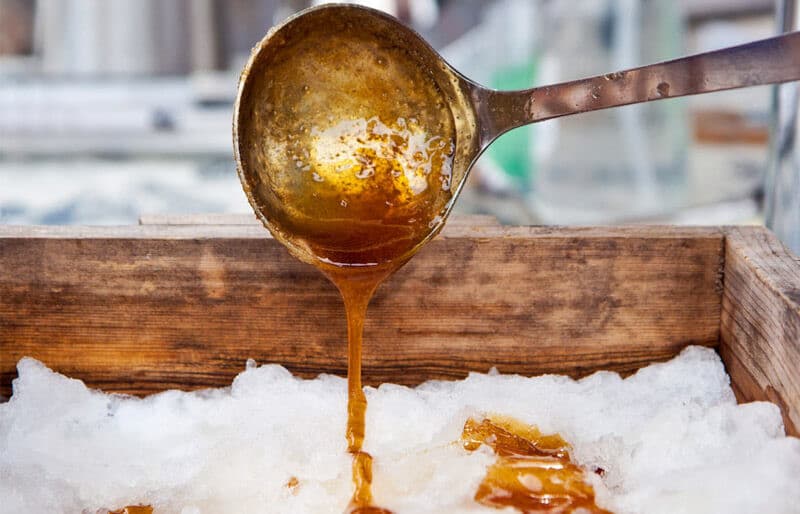 A metal ladle drips syrup onto a bed of snow.