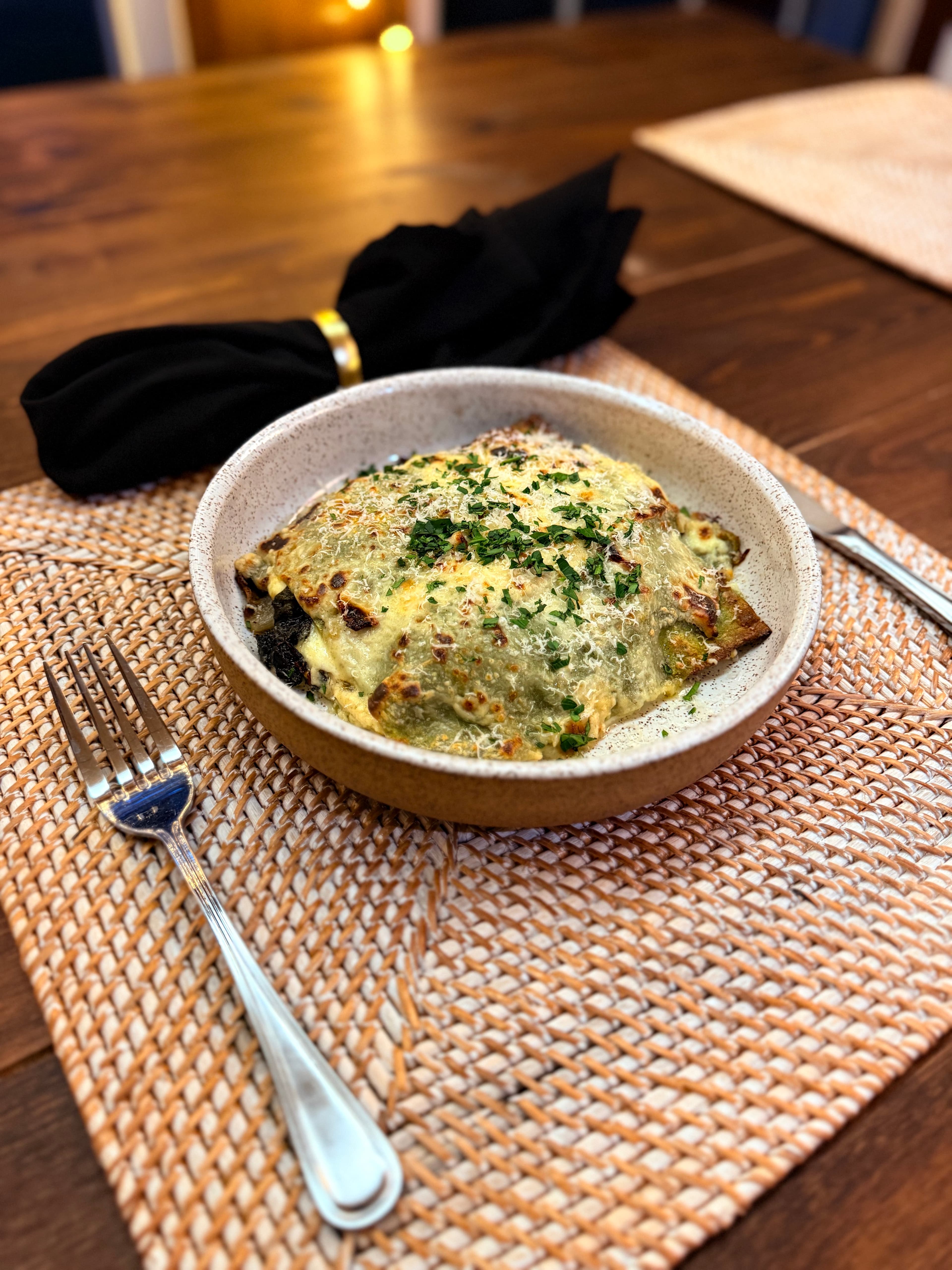 A creamy baked dish topped with cheese and herbs served in a bowl, next to a fork on a woven placemat.