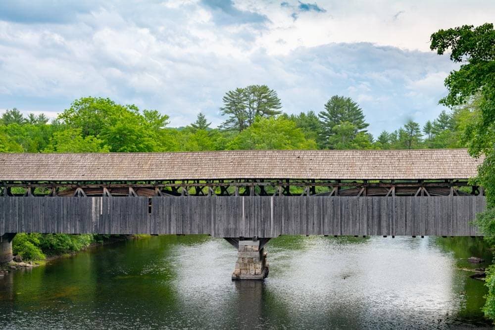 A wooden covered bridge spans a calm river surrounded by lush greenery.