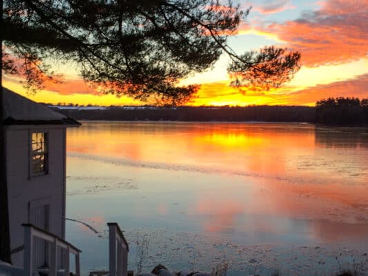 A serene sunset casts vibrant reflections on a calm lake, framed by a tree and a nearby cabin.