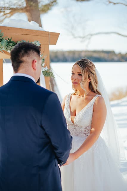 A bride and groom exchange vows under a wooden arch in a snowy landscape.