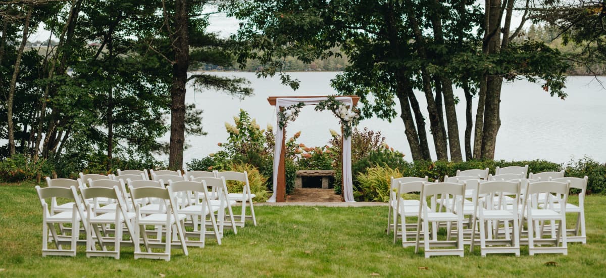 An outdoor wedding ceremony setup featuring white chairs facing a decorative arbor by a lake.