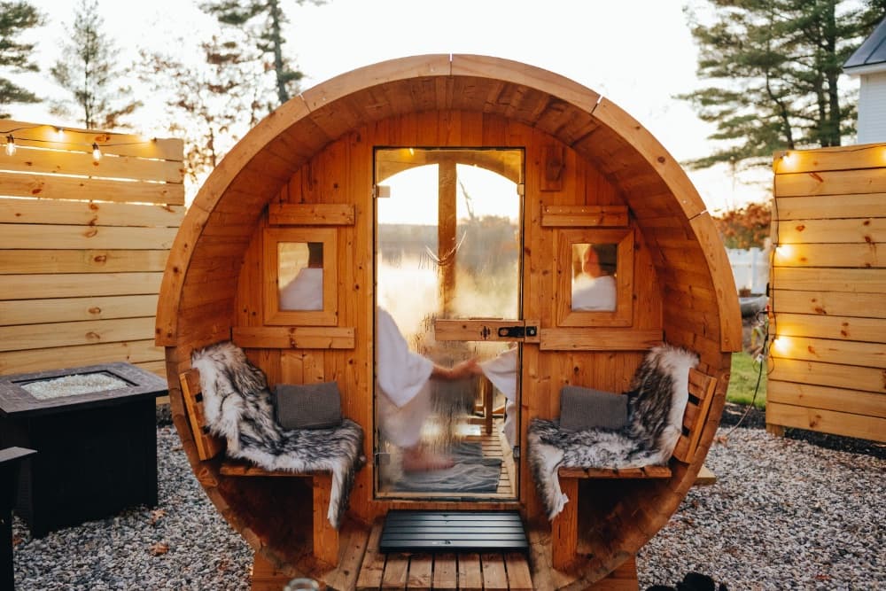 A wooden barrel sauna sits in a serene outdoor setting with plush seating and warm lighting.