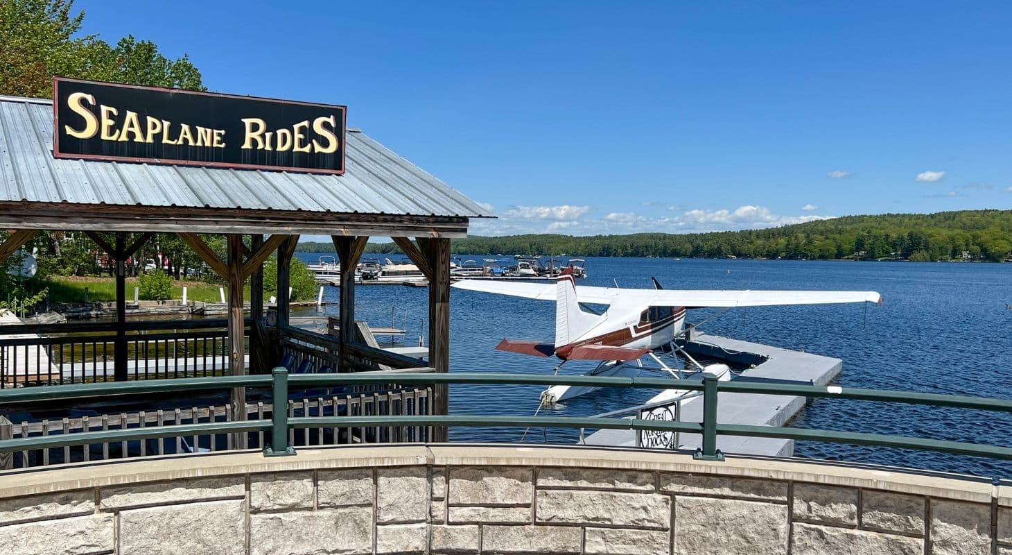 A seaplane is docked at a station labeled "Seaplane Rides" by a calm lake surrounded by greenery.
