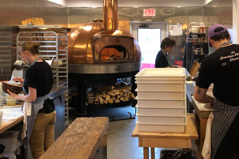 A bustling kitchen featuring two chefs preparing food beside a large copper pizza oven and stacks of ingredients.