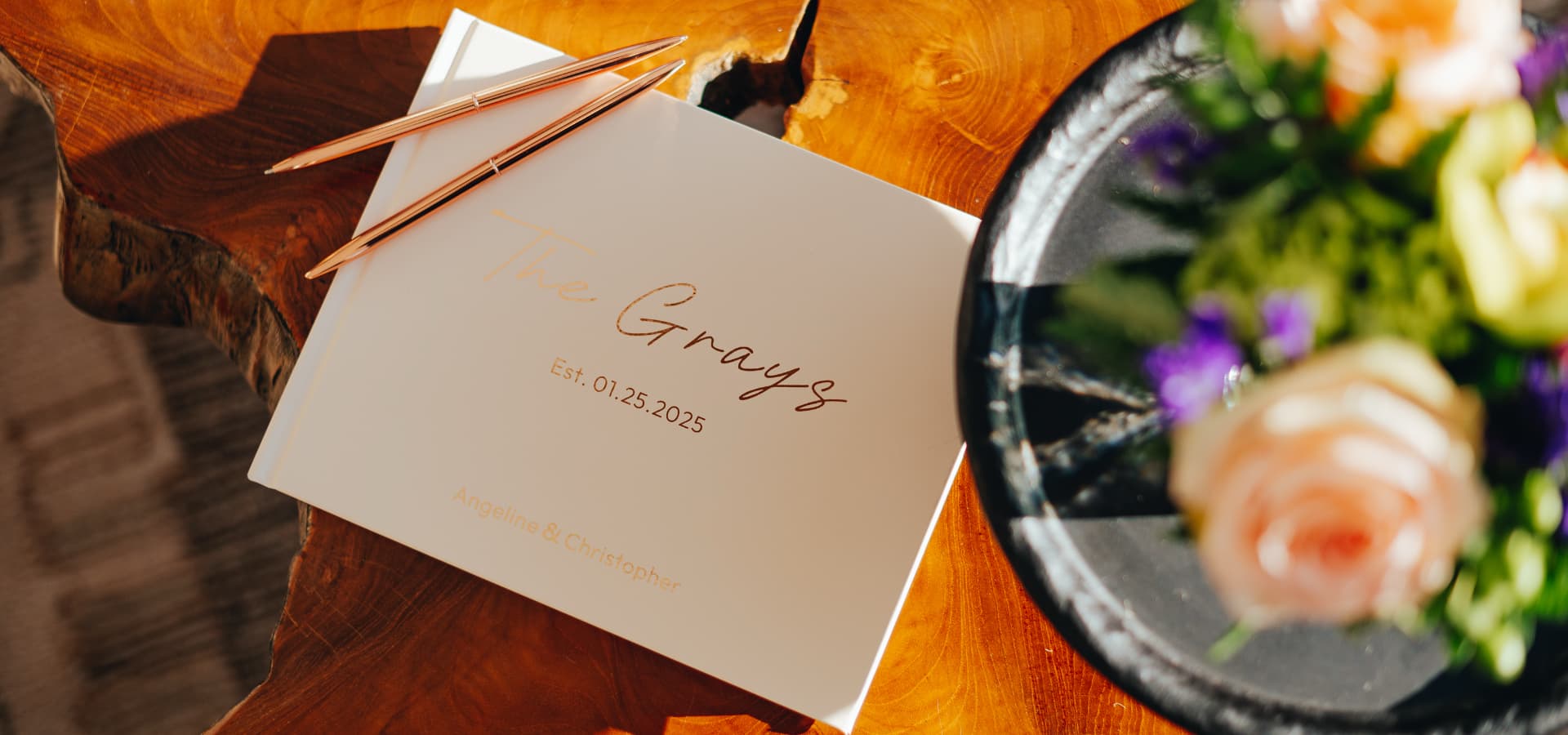 A wedding guest book titled "The Grays," established on January 25, 2025, lying on a wooden table beside two pens and a floral arrangement.