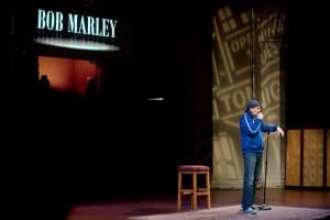 A comedian performs on stage with a spotlight and a stool, while a sign in the background reads "BOB MARLEY."