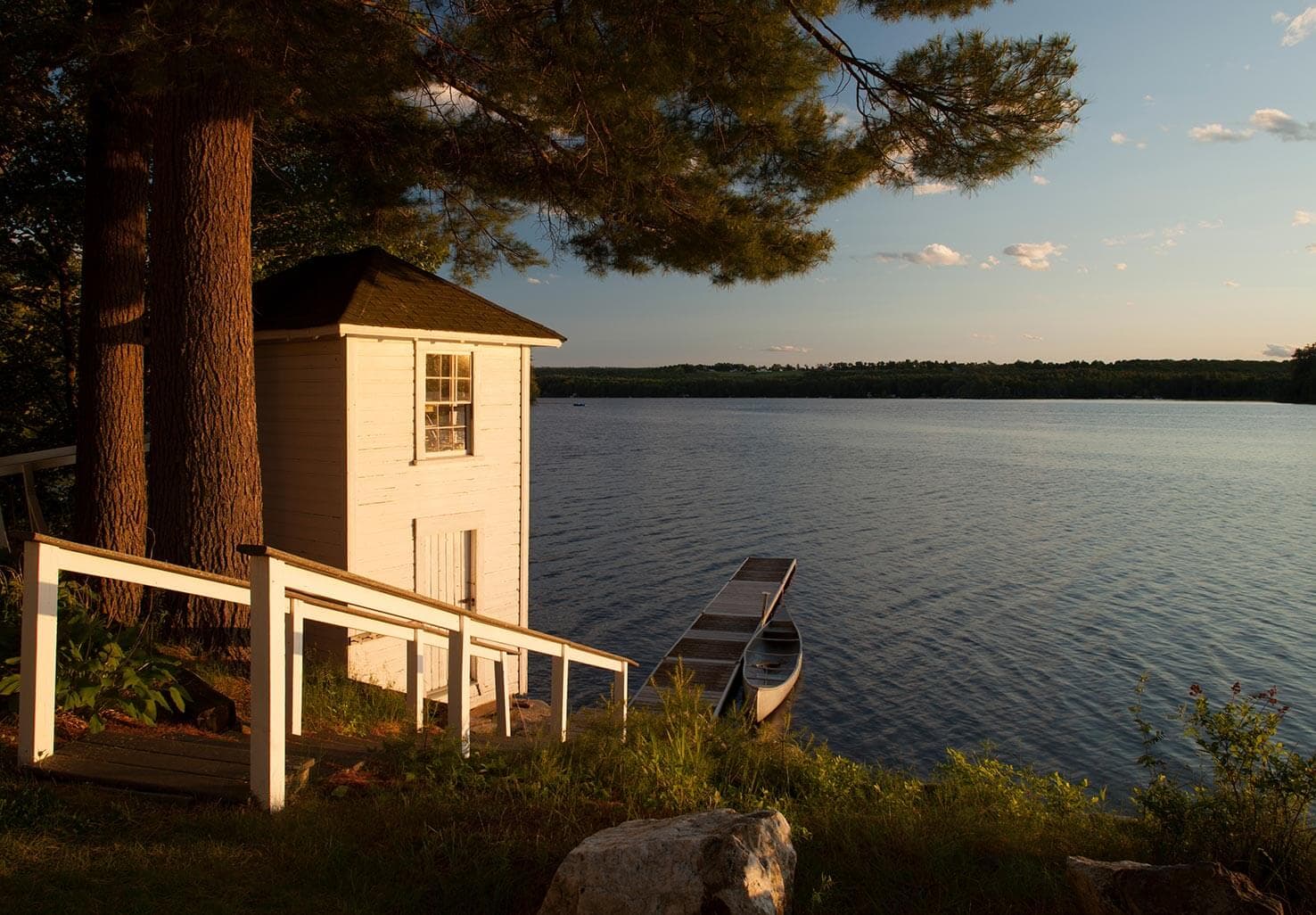 A serene lakeside scene featuring a small white boathouse, a canoe, and lush greenery under a sunset sky.