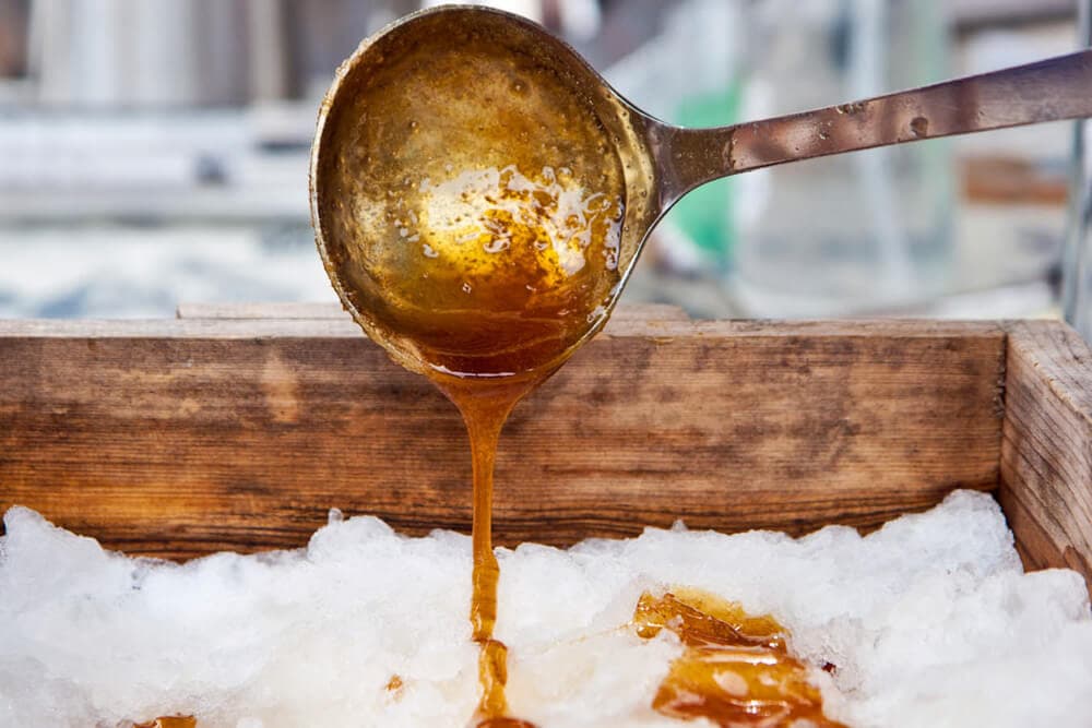 A brass ladle pours warm syrup over a bed of snow.