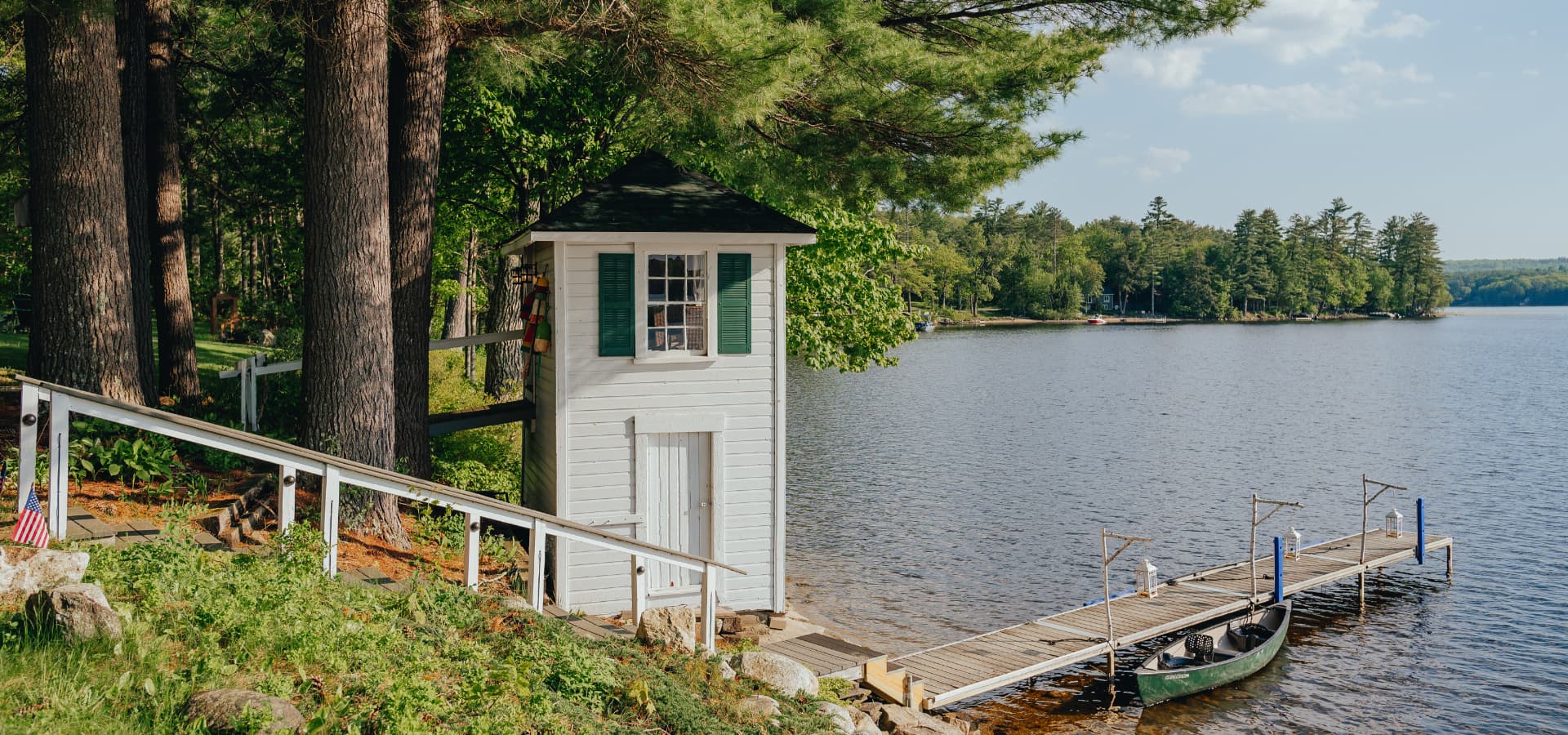 A small white building beside a serene lake and dock surrounded by trees.