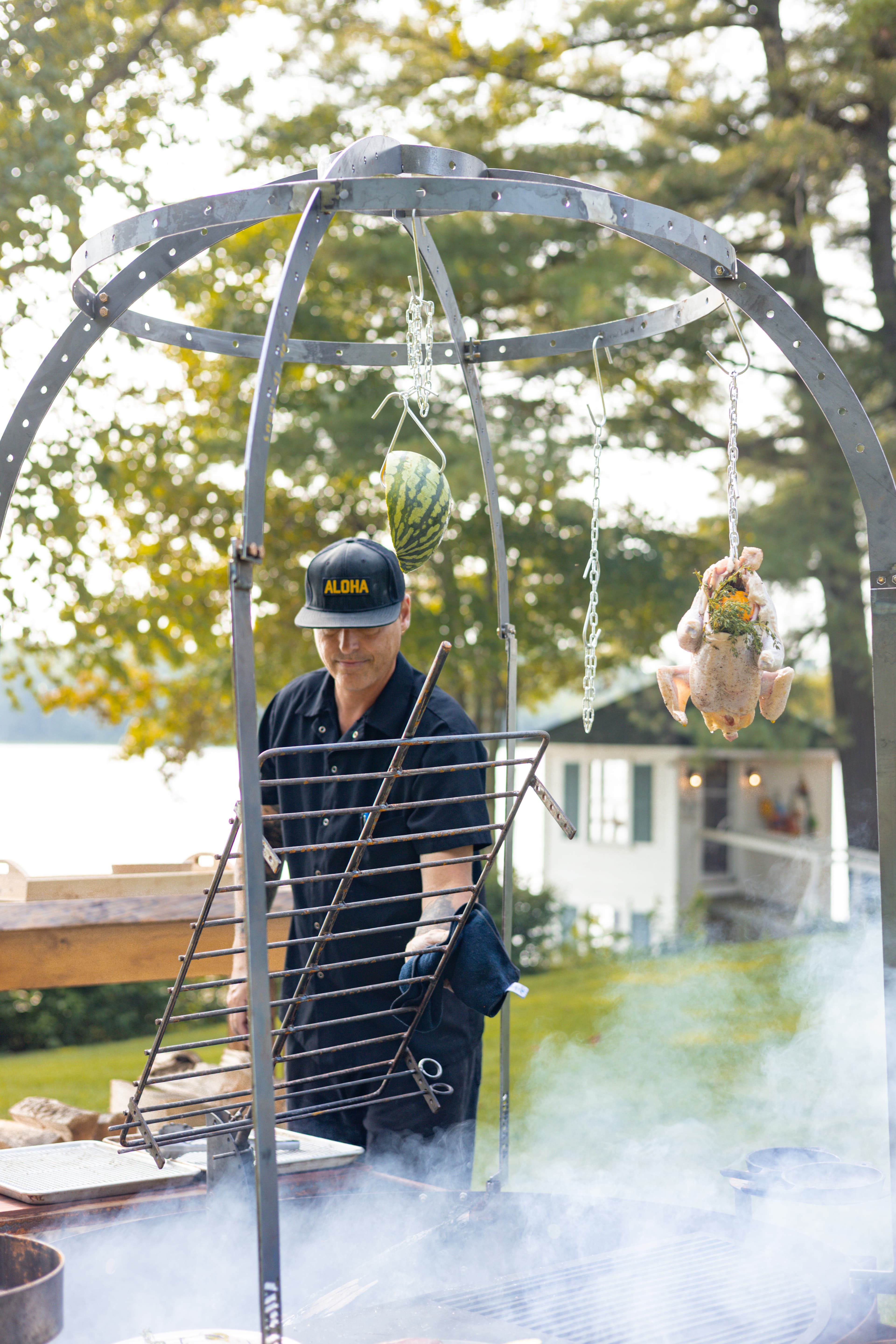 A man wearing a cap grills chicken and a watermelon over a smoky outdoor fire pit.