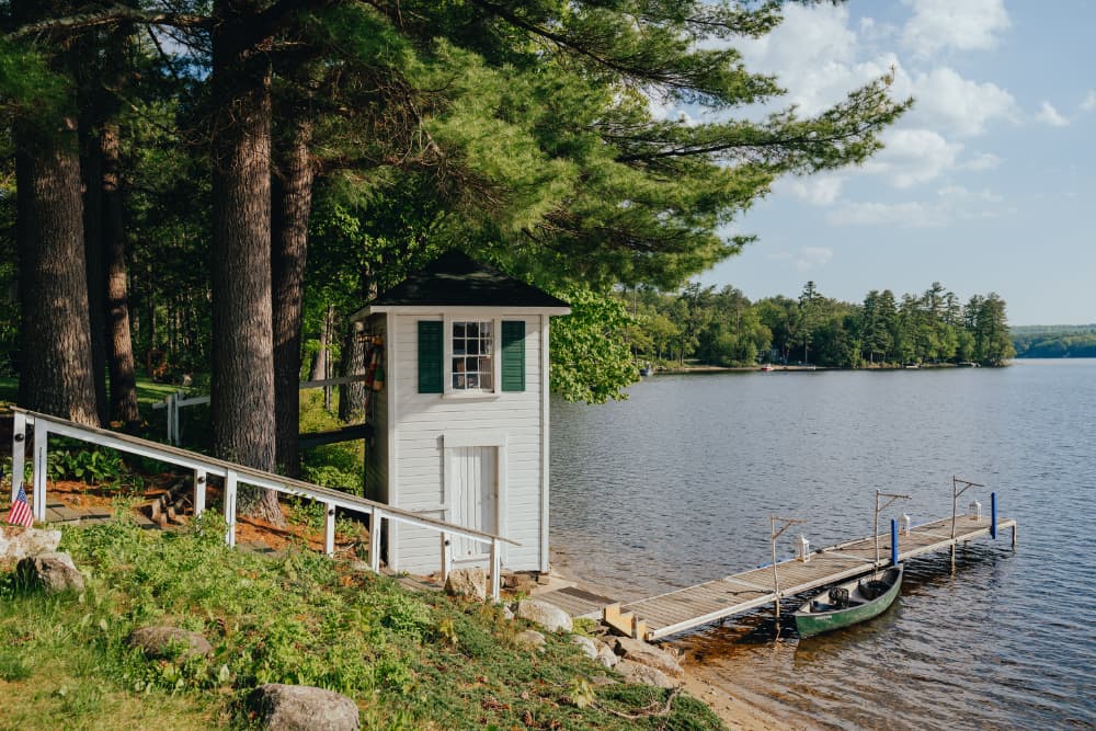 A serene lakeside view featuring a small white building and a dock with a canoe.