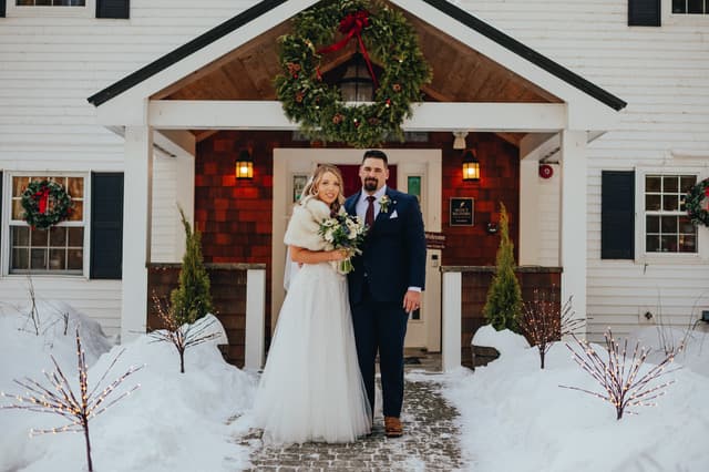 A bride and groom pose together outside a snow-covered venue decorated for the holidays.