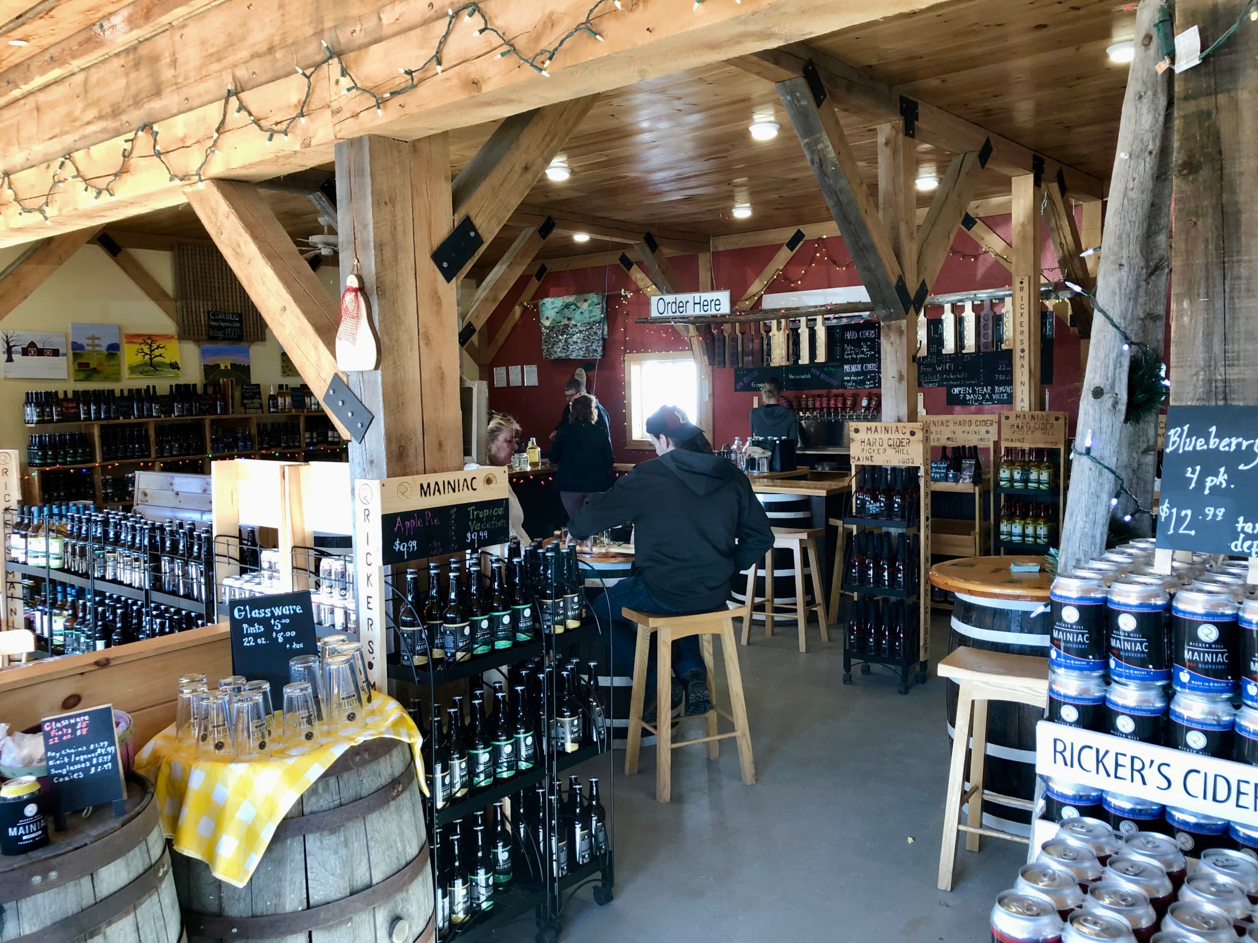 Interior of a rustic cider shop with wooden beams, shelves of bottled drinks, and customers seated at tables.