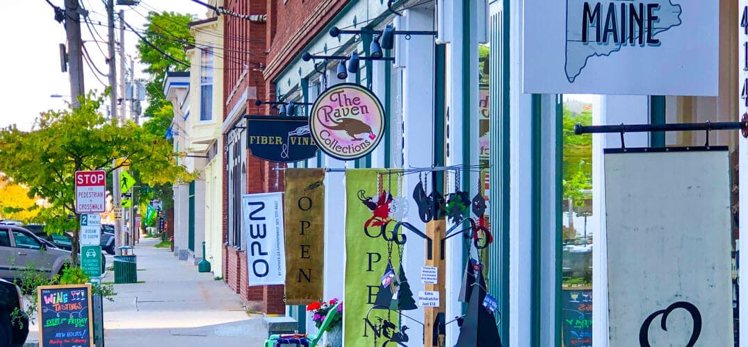 A vibrant street scene featuring storefronts with signs, including "The Raven Collection" and "OPEN," lined with trees in a charming coastal town.