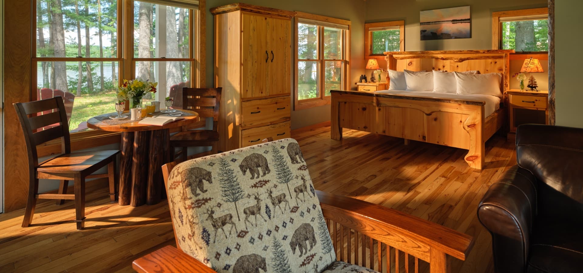 A cozy cabin interior featuring wooden furniture, a dining table with breakfast items, and large windows overlooking nature.