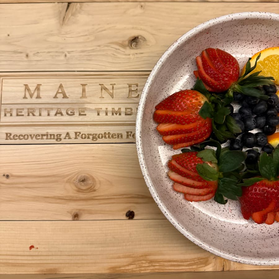 A bowl of fresh strawberries, blueberries, and an orange slice rests on a wooden surface with engraved text.