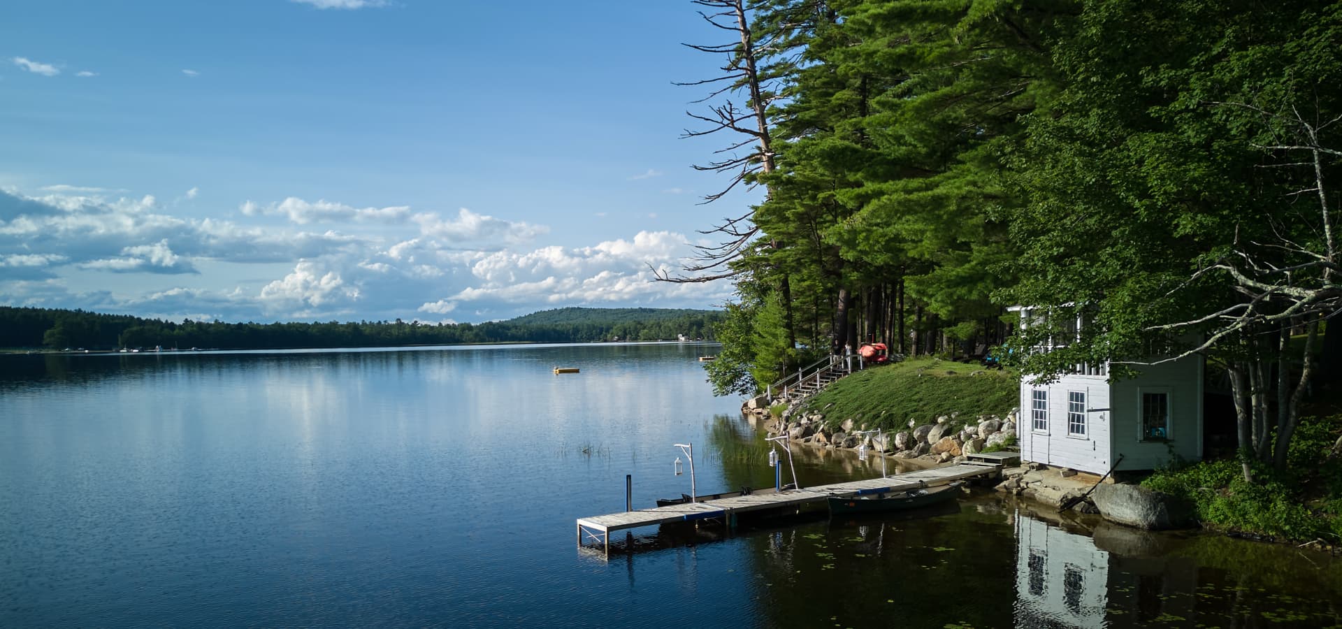 A tranquil lake scene with a white cabin, dock, and lush green pine trees under a blue sky.