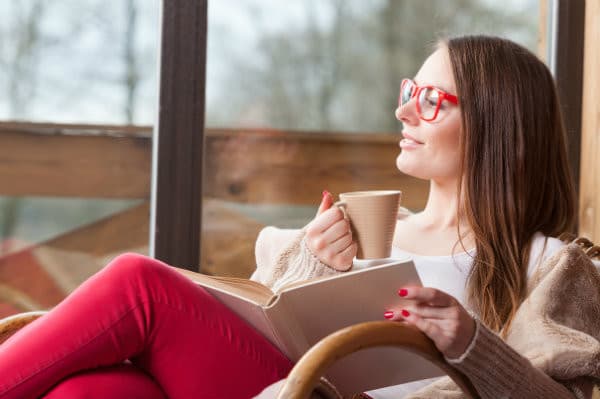 A woman in red glasses enjoys a cup of coffee while reading a book by a window.