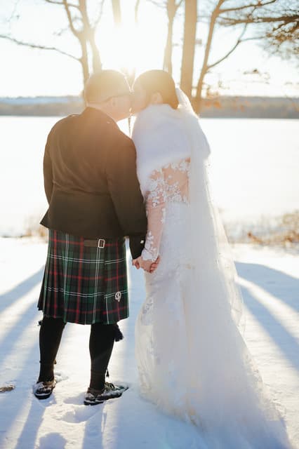 A couple in winter attire shares a kiss in a snowy landscape with sunlight shining behind them.