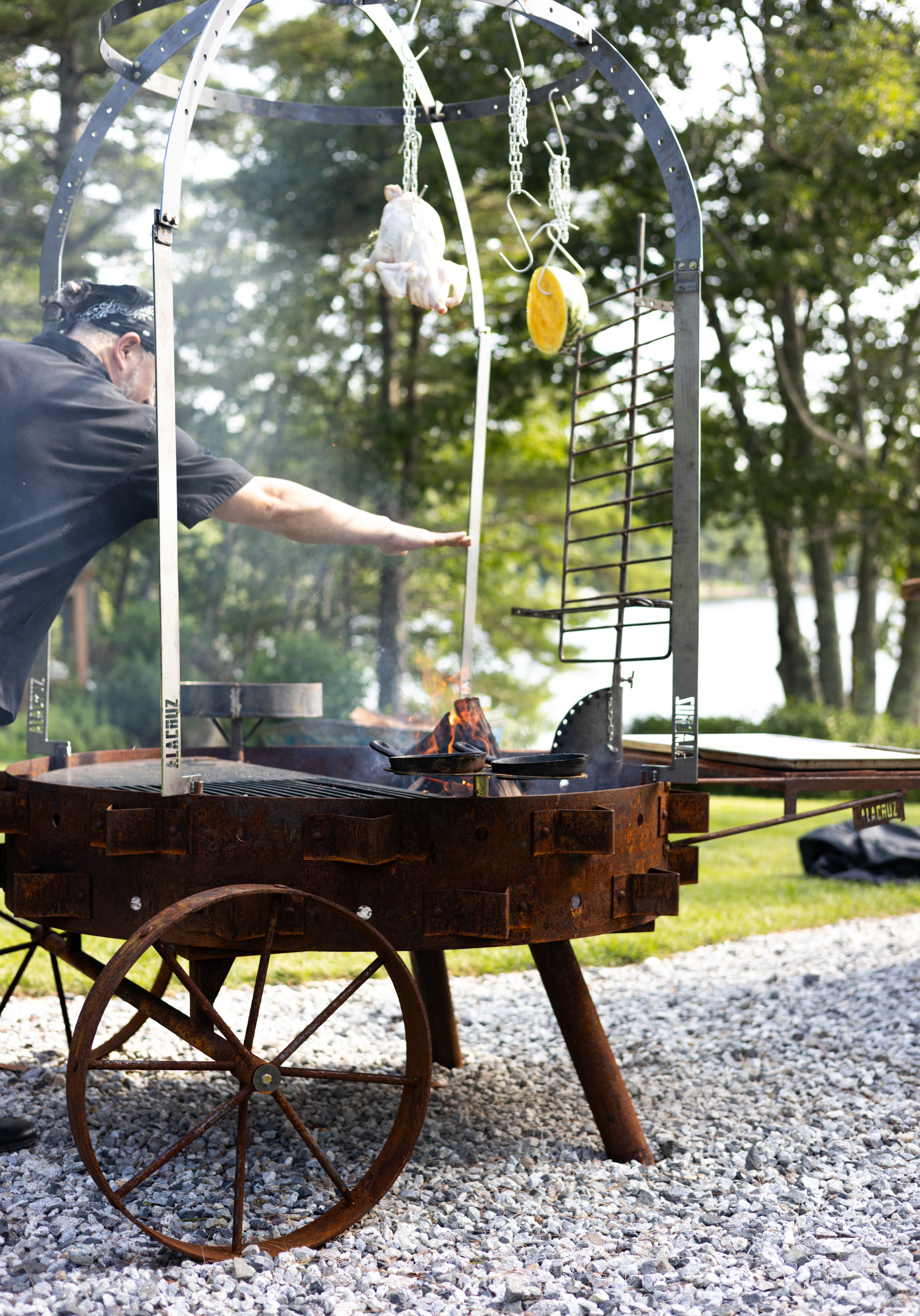 A person is grilling food over an open fire using a whimsical outdoor cooking apparatus on a gravel surface near a lake.