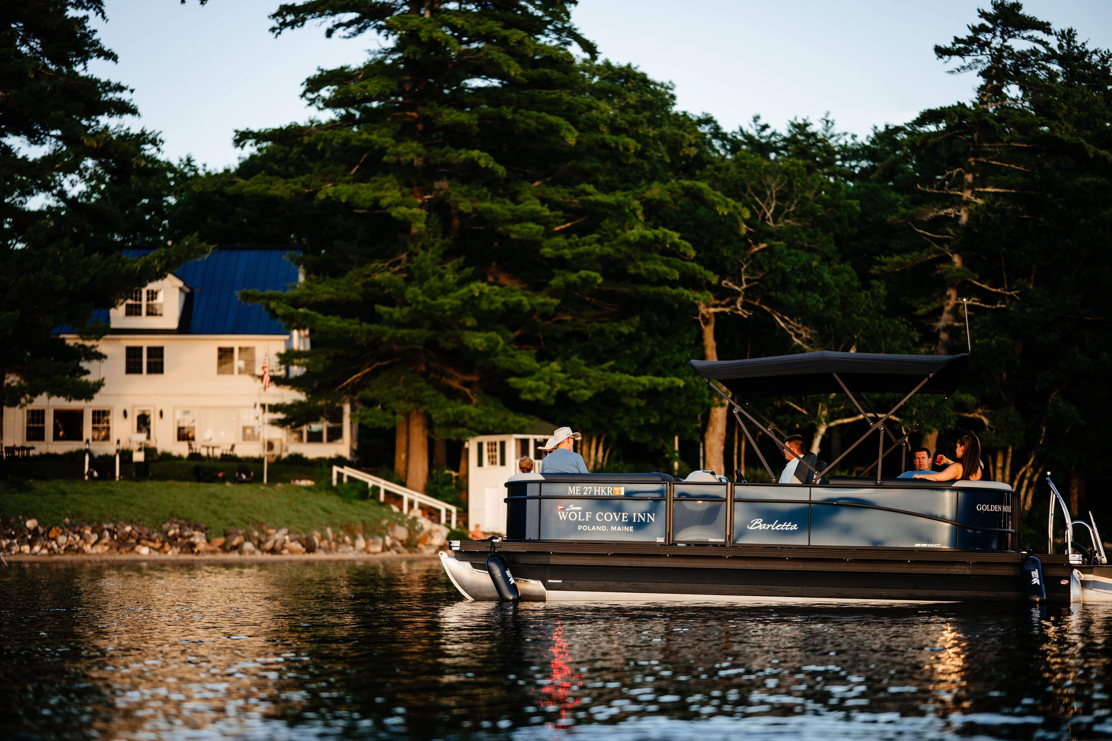 A pontoon boat with several people is seen on a calm lake near a lakeside inn surrounded by trees.