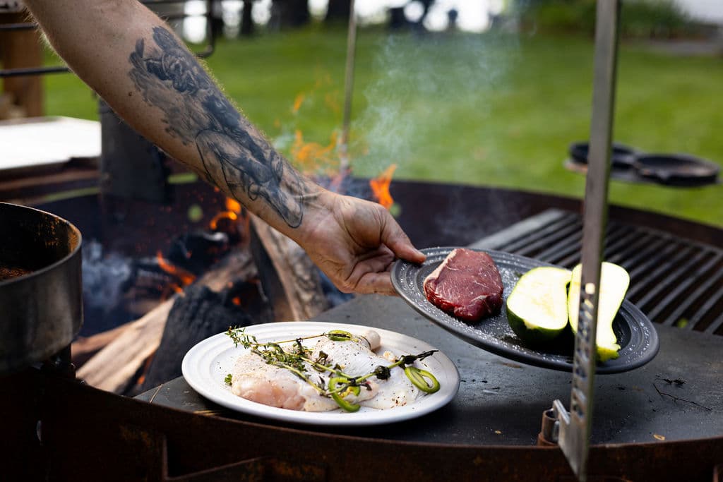 A tattooed hand holds a plate of meat and vegetables above a grill with an open flame.