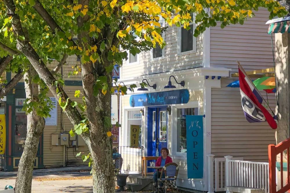 A woman sits outside a brightly colored restaurant adorned with flags and surrounded by autumn leaves.