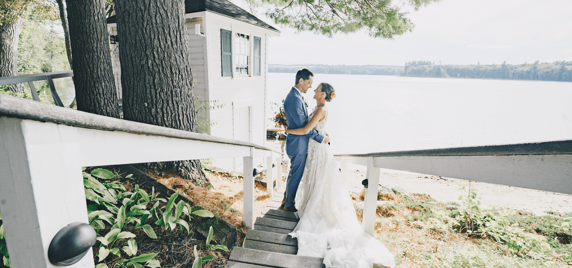 A couple in wedding attire embraces on a wooden staircase by a lakeside.