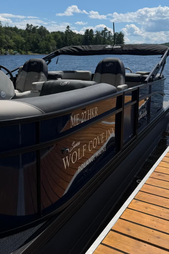 A dark blue boat named "Wolf Cove Inn" is docked at a wooden pier with a scenic lake in the background.