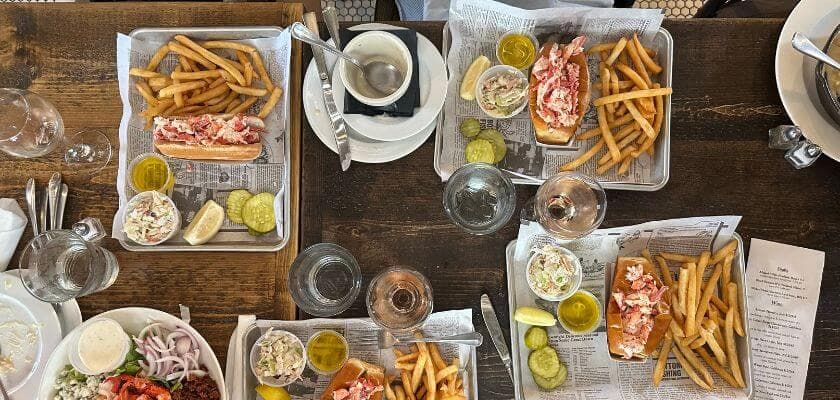 A wooden table spreads with lobster rolls, fries, pickles, and condiments, accompanied by glasses of water.