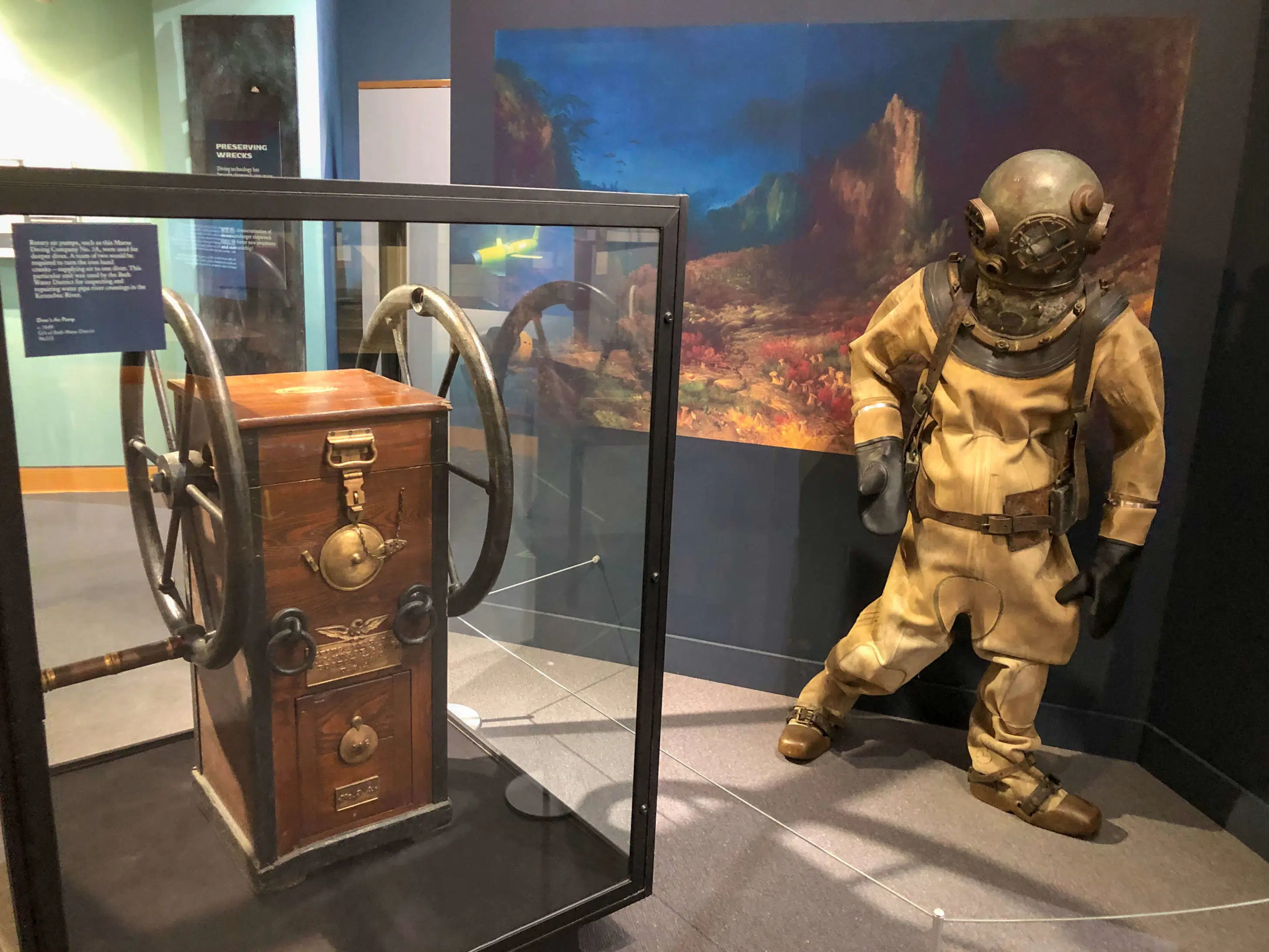 A vintage diving suit displayed next to a wooden dive control panel in a museum setting.