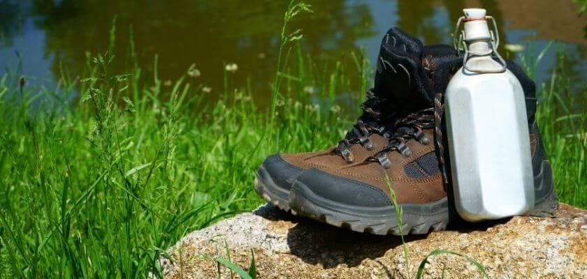 A pair of hiking boots next to a metal water bottle on a rock surrounded by grass.