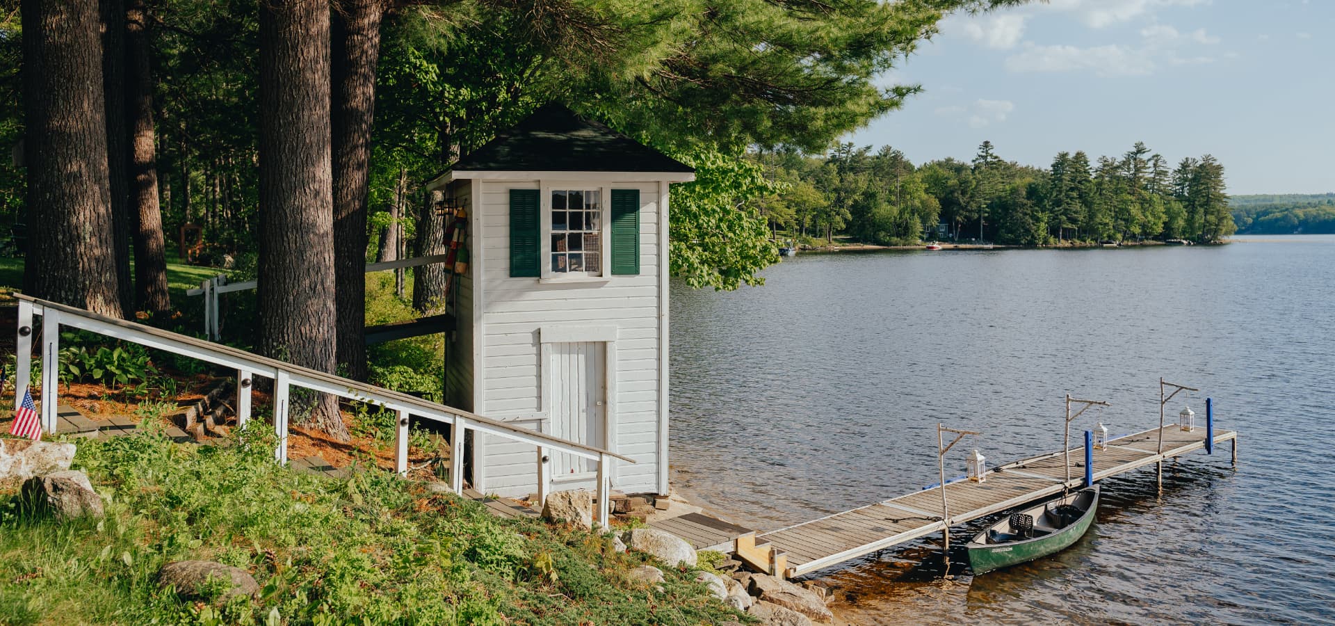 A small white building near a calm lake and wooden dock surrounded by trees.