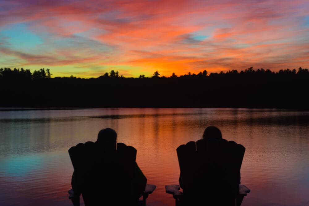 Two silhouettes sit in chairs by a lake, watching a colorful sunset.