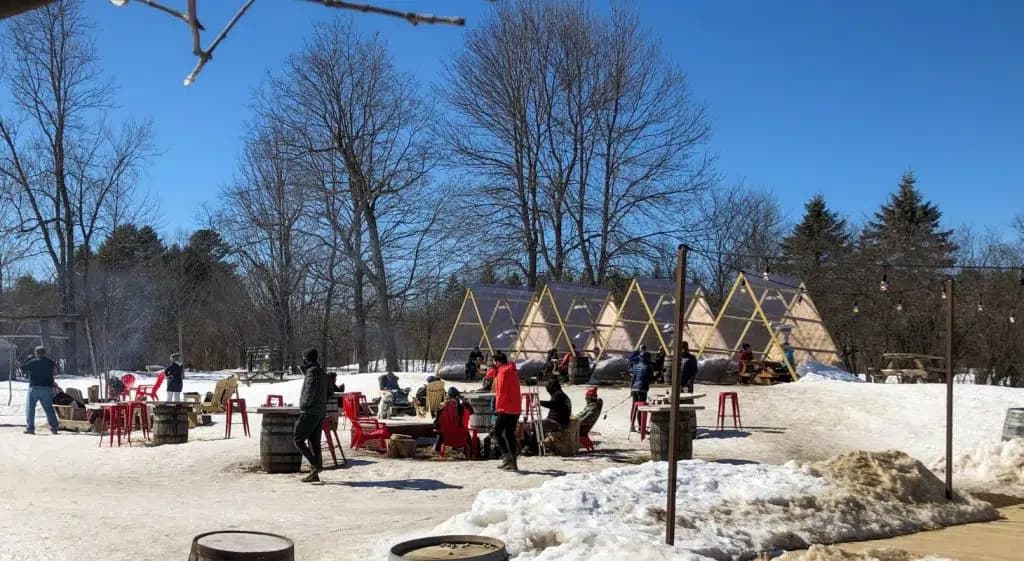 People enjoy a snowy outdoor gathering near teepee-like structures on a sunny day.