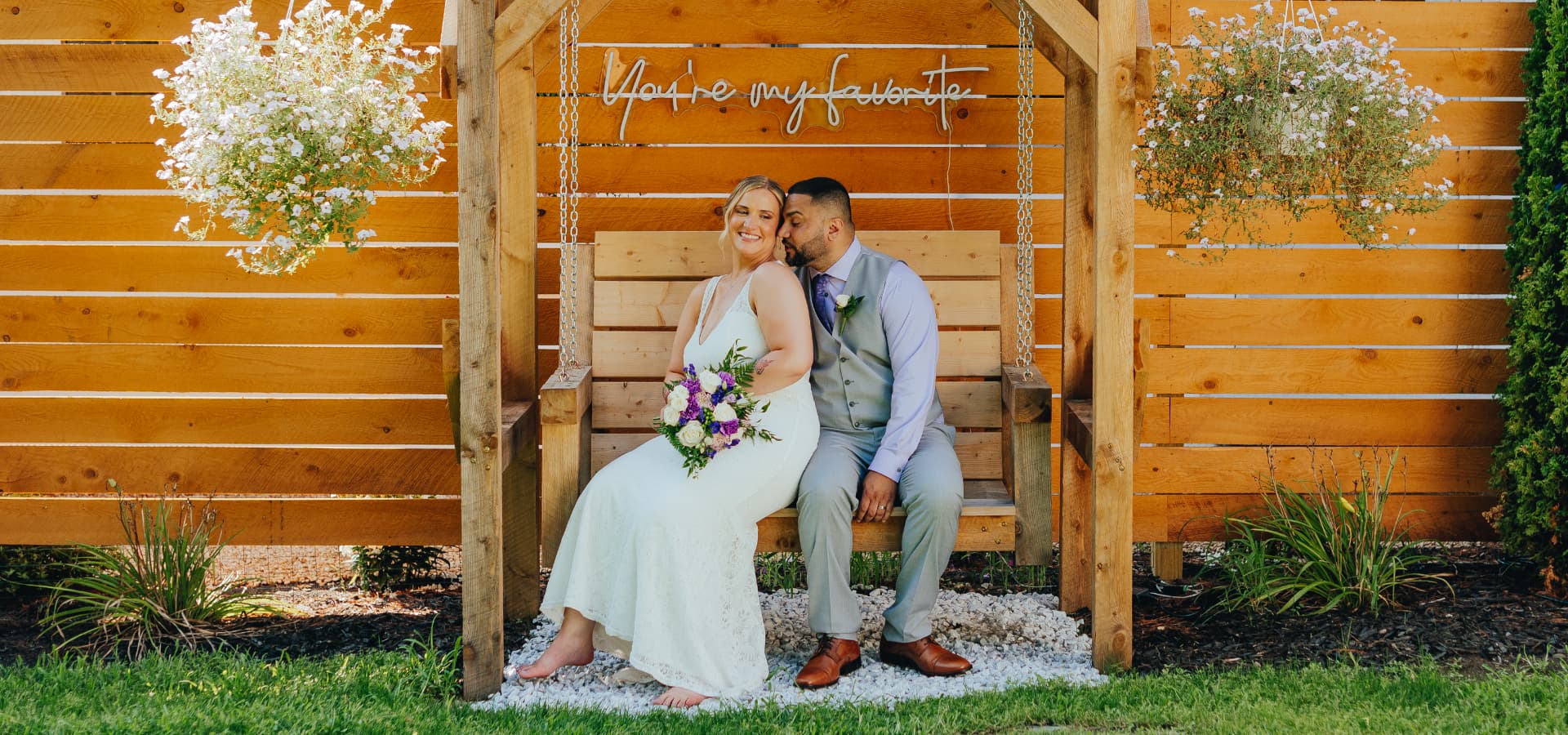 A happy couple sits on a wooden swing surrounded by flowers, celebrating their love at a wedding.