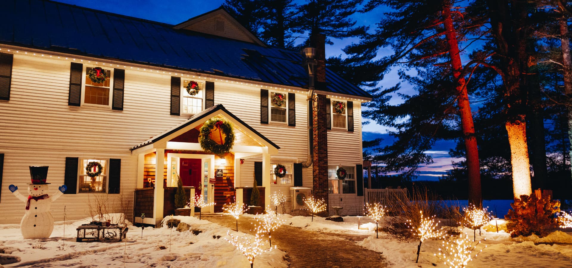 A festive home illuminated by holiday lights and a snowman in a snowy landscape at dusk.