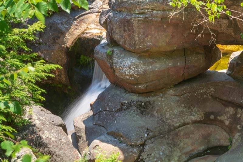 A small waterfall flows between large, moss-covered rocks surrounded by greenery.